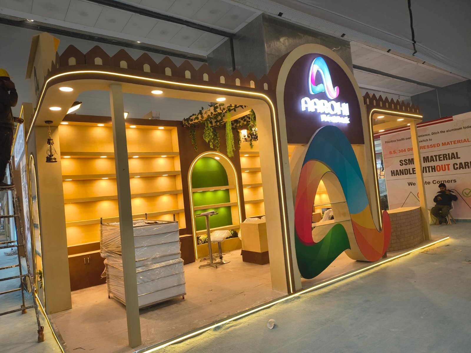 Aarohi exhibition stall during build-up, with arched backlit shelving and a curved logo feature