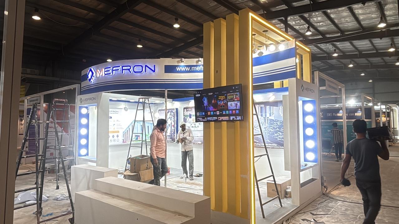 Mefron exhibition stall during setup, with a curved illuminated fascia and a yellow framed entrance